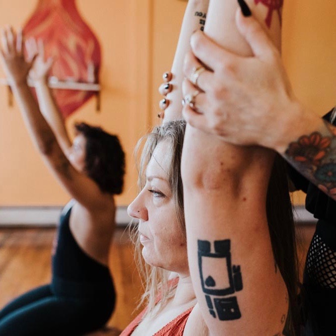 A woman with a prominent tattoo on her arm is aided in stretching her arms above her head.