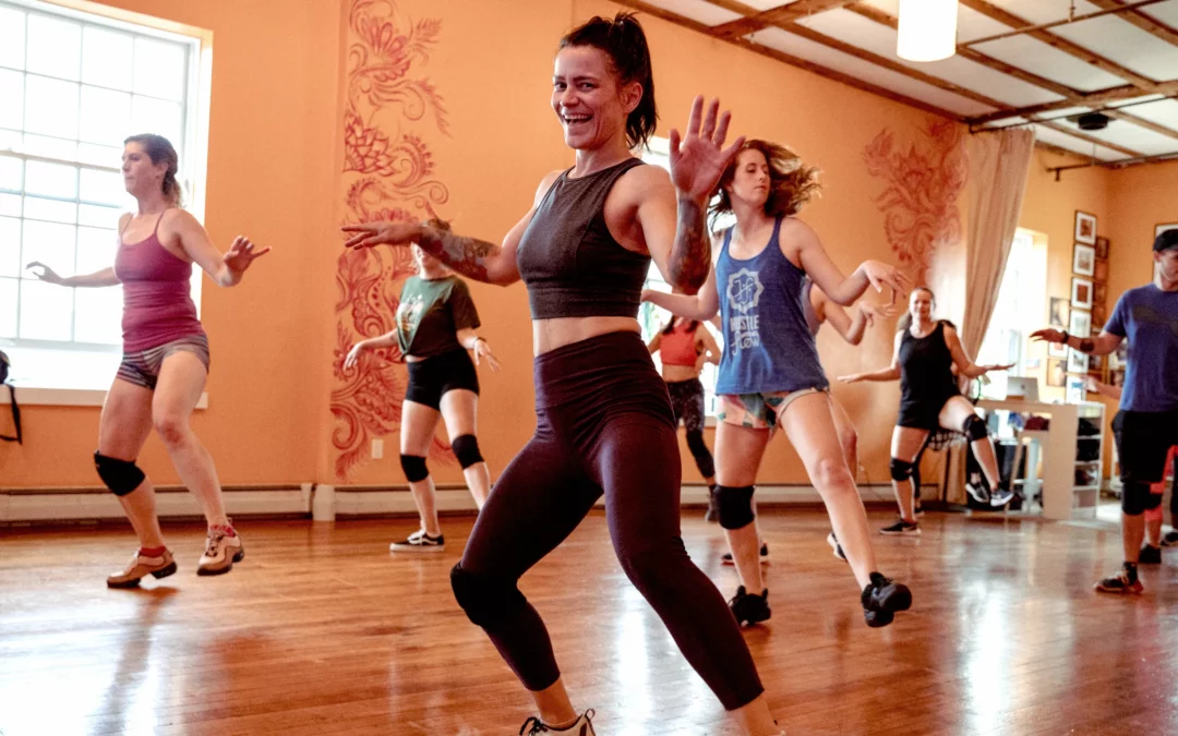 A lively dance class in session at a studio with wooden floors. The instructor is leading the class with a big smile. Participants are actively following along, enjoying the energetic routine.