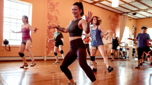 A lively dance class in session at a studio with wooden floors. The instructor is leading the class with a big smile. Participants are actively following along, enjoying the energetic routine.