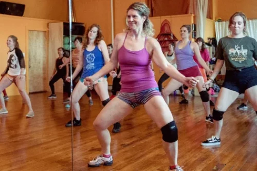 A group of women participating in a dance class at a studio. The woman in the foreground is wearing a pink tank top and striped shorts. The participants follow along with focused expressions.
