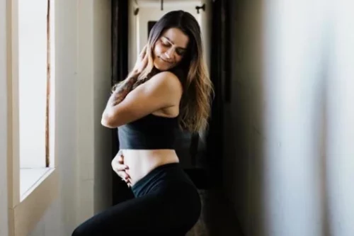 A woman dressed in black activewear poses confidently in a narrow hallway. She stands sideways, with one hand on her chest and the other on her hip, looking down with a smile.