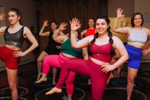 A group of women in colorful workout attire smiling and exercising on mini trampolines in a fitness studio. They are raising one arm and lifting one knee.