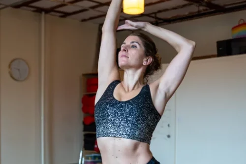 A woman in a sparkly black sports bra raising her right arm above her head while looking upward.