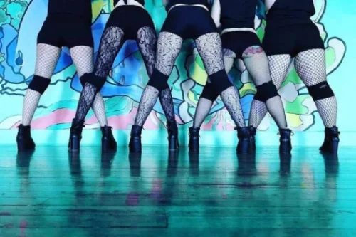 A group of women dressed in black shorts, fishnet stockings, and knee pads face the wall, posing in front of a colorful mural, showcasing their high heels. The photo only shows the women from the waist down.