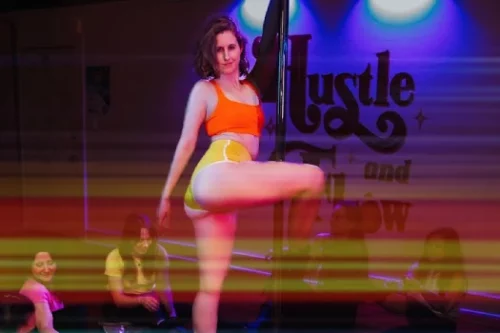A woman in an orange top and yellow shorts performs a pole dance move in a studio with purple lighting. She holds the pole with one hand and strikes a confident pose. Yellow light streaks across the photo.