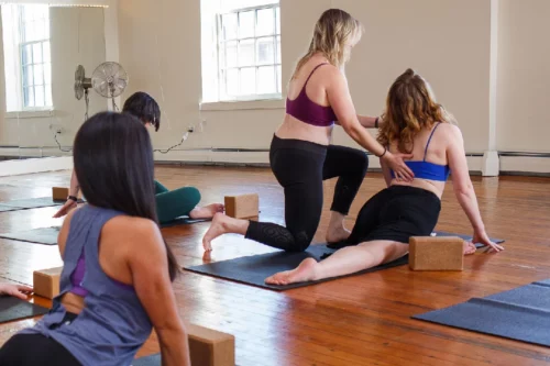 A group of women in a yoga studio on yoga mats with yoga blocks. One woman, wearing a burgundy top, assists another woman in a blue top by gently adjusting her posture.