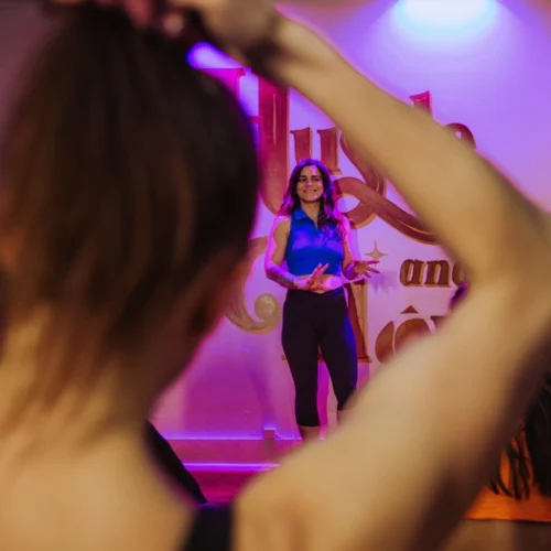 During a class, a participant in the foreground is tying her hair back, with the focus of the photo on the female instructor smiling and gesturing in the background.