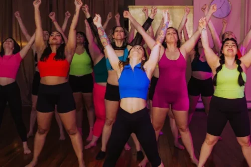 A group of women participating in an energetic dance workout. They are standing with their arms raised in the air, wearing colorful workout outfits, and engaging enthusiastically in the activity.