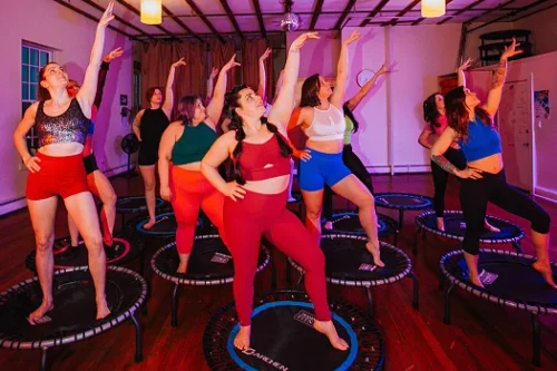 A group of women participating in a trampoline workout class at Hustle and Flow studio. They are standing on mini trampolines, wearing colorful workout outfits, and striking a pose with one arm raised.