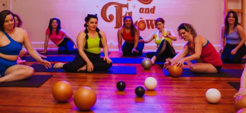 A group of women participating in a fitness class at Hustle and Flow studio. They are seated on yoga mats with exercise balls of various colors around them.