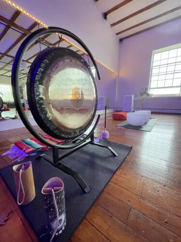 Sound-Bath-Elixir-Venus-Gong-Hustle-and-Flow-1.jpg A serene woman receiving a Reiki treatment at Hustle and Flow studio. She is lying down on a colorful striped blanket with her eyes closed, wearing a tank top with a tiger graphic and denim jeans. The practitioner's hands are gently placed on her head.