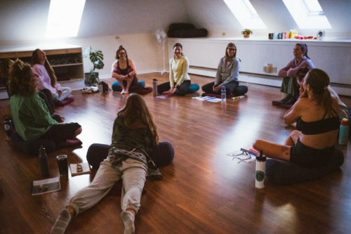 Portland-Maine-Hustle-and-Flow-Dance-Cardio-Teacher-Training-3 A woman dressed in black activewear poses confidently in a narrow hallway. She stands sideways, with one hand on her chest and the other on her hip, looking down with a smile.