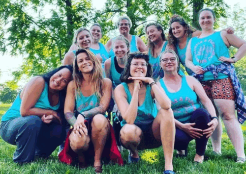 A group of women dressed in black shorts, fishnet stockings, and knee pads face the wall, posing in front of a colorful mural, showcasing their high heels. The photo only shows the women from the waist down.