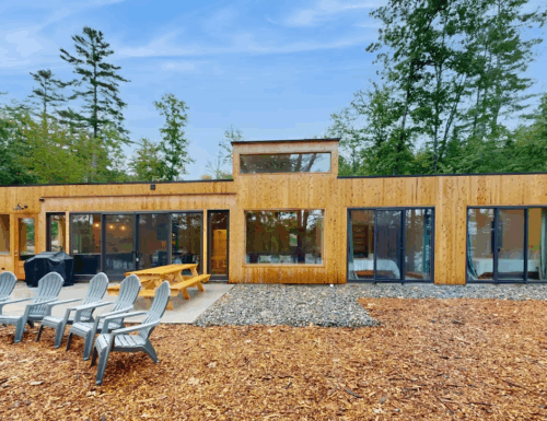 A woman dressed in black activewear poses confidently in a narrow hallway. She stands sideways, with one hand on her chest and the other on her hip, looking down with a smile.